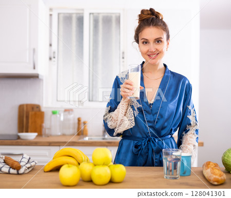 Smiling young woman in blue robe drinking milk in home kitchen 128041301