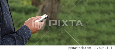 Outside in nature, a male hand interacts with a smartphone, its green background complementing the natural setting. Perfect for a copy space design Outside in nature, a male hand interacts with a smartphone, its green background complementing the natural setting. Perfect for a copy space design 128042101