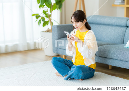 在房間裡操作智慧型手機的年輕女子 在房間裡操作智慧型手機的年輕女子 128042687