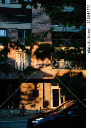 Vertical screen: sunlight illuminating brick facade of modern residential building at sunset with bicycles parked on sidewalk and car passing by, seen through foliage. Brick apartment exterior Vertical screen: sunlight illuminating brick facade of modern residential building at sunset with bicycles parked on sidewalk and car passing by, seen through foliage. Brick apartment exterior 128044791