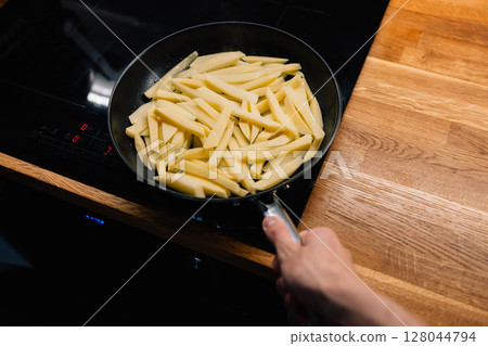 First-person view: man holding handle of pan filled with raw French fries on sleek black vitroceramic stove in modern home kitchen, emphasizing healthy food preparation concept. Cooking French fries 128044794