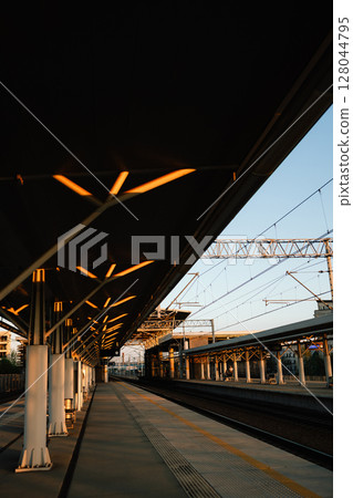 Vertical screen: modern architecture of empty train station platform with illuminated ceiling and visible overhead lines at sunset, creating peaceful and captivating urban scene. Minimalist station Vertical screen: modern architecture of empty train station platform with illuminated ceiling and visible overhead lines at sunset, creating peaceful and captivating urban scene. Minimalist station 128044795