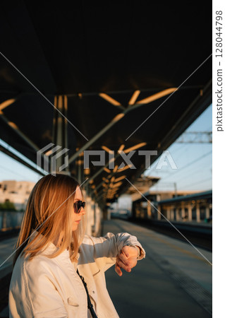 Vertical screen: blonde woman executive wearing sunglasses and white blazer checking wristwatch while waiting near railway tracks during golden sunset lighting. Woman checking time, waiting for train Vertical screen: blonde woman executive wearing sunglasses and white blazer checking wristwatch while waiting near railway tracks during golden sunset lighting. Woman checking time, waiting for train 128044798