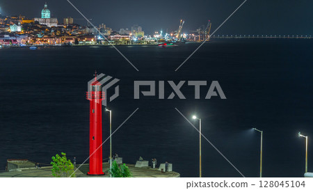 Aerial night timelapse of Cacilhas red lighthouse near bus and railway station with the Tagus River, Lisbon glowing skyline. 128045104