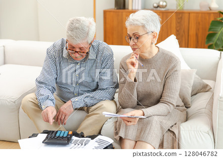 Middle aged senior couple sit with laptop and paper document. Older mature man woman reading paper bill pay online at home managing bank finances calculating taxes planning loan debt pension payment 128046782