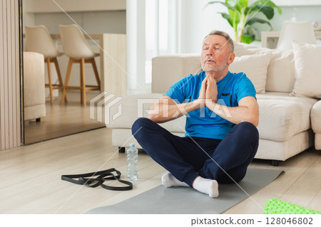 Yoga mindfulness meditation. Senior adult mature man practicing yoga at home. Mid age old grandfather sitting in lotus pose on yoga mat meditating relaxing. Older man doing breathing practice Yoga mindfulness meditation. Senior adult mature man practicing yoga at home. Mid age old grandfather sitting in lotus pose on yoga mat meditating relaxing. Older man doing breathing practice 128046802