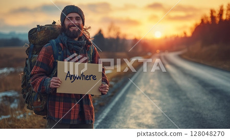 A happy hiker with a backpack holds a sign saying 'Anywhere,' signifying his journey and freedom on an open road at sunset 128048270