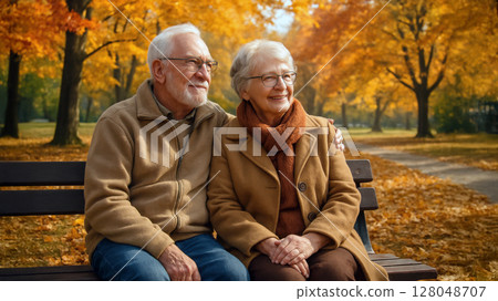 Senior couple sitting on park bench surrounded by autumn trees with colorful leaves, smiling and enjoying peaceful fall day together. Warm emotional scene. 128048707