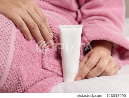 Woman in pink bathrobe holding in hand blank cream tube on white bedsheet closeup, cosmetic mockup Woman in pink bathrobe holding in hand blank cream tube on white bedsheet closeup, cosmetic mockup 128052090