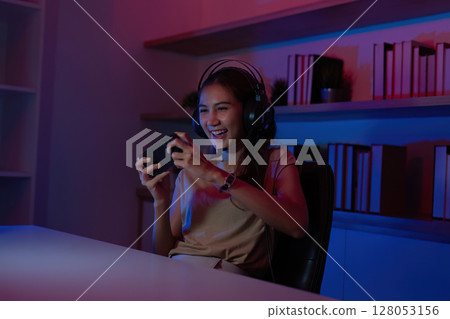 Gaming and Streaming. Joyful young woman playing mobile game with headset in colorful lighting. Gaming and Streaming. Joyful young woman playing mobile game with headset in colorful lighting. 128053156