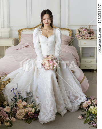 Woman in a white wedding dress sits on a bed with a bouquet of flowers in her 128053507