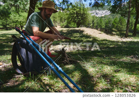 Man Preparing Bonfire in Forest Clearing with Backpack and Hiking Equipment 128053826