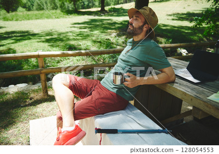 Man Relaxing Outdoors Near Wooden Table with Laptop and Cup in Summer Setting 128054553
