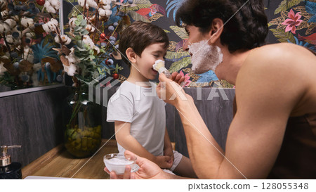 Father playfully applies shaving cream to young son's nose in colorful bathroom setting Father playfully applies shaving cream to young son's nose in colorful bathroom setting 128055348