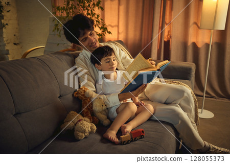 Father reading bedtime story to son on cozy sofa with warm lighting and toys nearby 128055373