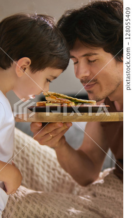 Family moments. Father and son enjoying breakfast in bed with sandwich on Fathers Day morning Family moments. Father and son enjoying breakfast in bed with sandwich on Fathers Day morning 128055409