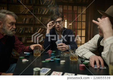Male friends company playing cards in a dimly lit bar while sipping on drinks 128055539