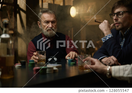An experienced mature man sits at the poker table enjoying his whiskey and playing with chips 128055547