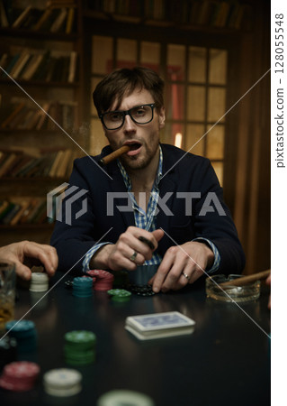 Man holding a cigar at a poker table with alcohol drink nearby 128055548