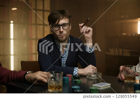 Man holding a cigar at a poker table with alcohol drink nearby 128055549