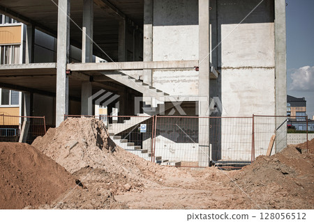 Construction site showing a modern building under progress with exposed concrete and sand piles during daylight Construction site showing a modern building under progress with exposed concrete and sand piles during daylight 128056512