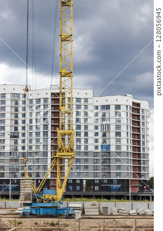 A large tower crane at a construction site against the backdrop of a modern monolithic house. Modern housing construction. industrial engineering. Construction of mortgage housing. A large tower crane at a construction site against the backdrop of a modern monolithic house. Modern housing construction. industrial engineering. Construction of mortgage housing. 128056945