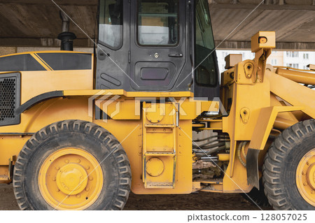 Heavy wheel loader with a bucket at a construction site. Close-up. Equipment for earthworks, transportation and loading of bulk materials - earth, sand, crushed stone. Heavy wheel loader with a bucket at a construction site. Close-up. Equipment for earthworks, transportation and loading of bulk materials - earth, sand, crushed stone. 128057025