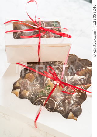 A hand holds a beautifully packaged holiday cookie box, tied with a vibrant red ribbon. Inside, assorted decorated gingerbread cookies peek through the clear window, making this a perfect edible gift A hand holds a beautifully packaged holiday cookie box, tied with a vibrant red ribbon. Inside, assorted decorated gingerbread cookies peek through the clear window, making this a perfect edible gift 128058240