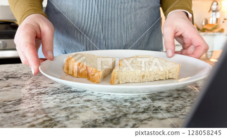 A person in a striped apron places cut pieces of bread onto a white plate on a marble countertop. The kitchen scene is clean and bright. 128058245