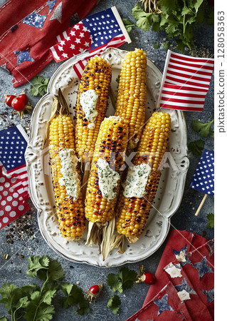 Bright summer flat lay of grilled corn on the cob brushed with herbed butter and sprinkled with cotija cheese, arranged on a white platter surrounded by mini American flags, red napkins, and fresh 128058363