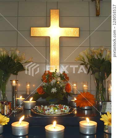 A lit candle is on a table in front of a cross 128058517