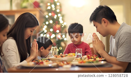 A family of four is praying at a table with a Christmas tree in the background 128058728