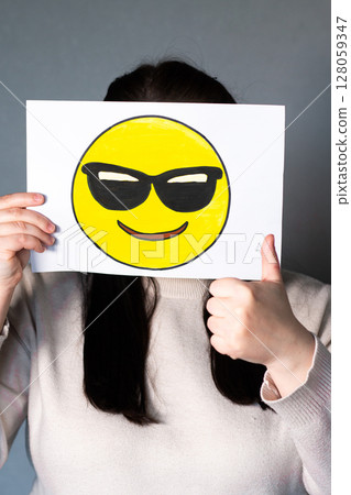 Woman holding happy emoji with sunglasses mask in front of face. Making thumbs up. World emoji day 17 July. 128059347