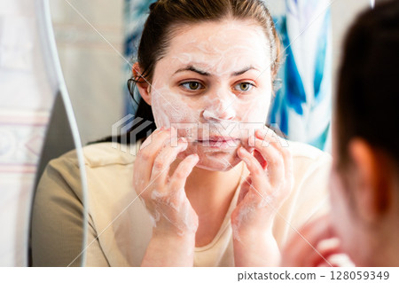 Woman removing makeup with face cleansing foam. Reflection in bathroom mirror. Beauty routine. Dermatology concept. 128059349