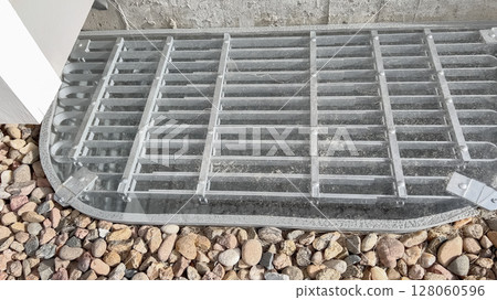 A metal grate cover is installed over a basement window well filled with natural gravel. The cover is flush with the concrete foundation wall. A metal grate cover is installed over a basement window well filled with natural gravel. The cover is flush with the concrete foundation wall. 128060596