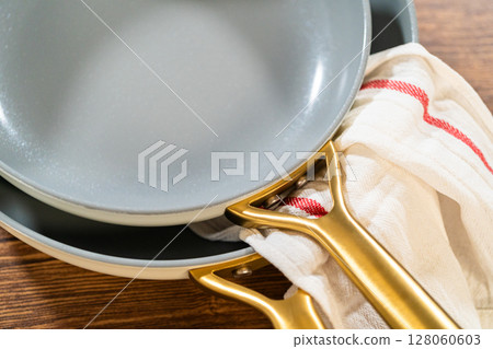 A set of stacked ceramic frying pans with matte gold handles, resting on a wooden surface and accented by a folded kitchen towel. The image showcases modern and coordinated cookware. A set of stacked ceramic frying pans with matte gold handles, resting on a wooden surface and accented by a folded kitchen towel. The image showcases modern and coordinated cookware. 128060603