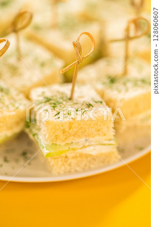 Delicate mini cucumber sandwiches made with fluffy white bread, a layer of herbed cream cheese, and thinly sliced cucumbers. Each square is topped with a sprinkle of fresh dill and secured with a 128060607