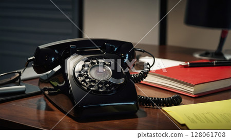 Black rotary phone sitting on wooden desk with documents and notebook 128061708
