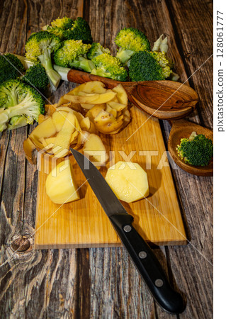 A sharp knife lays on a wooden cutting board with peeled and cut potatoes with broccoli florets on a wooden surface. A sharp knife lays on a wooden cutting board with peeled and cut potatoes with broccoli florets on a wooden surface. 128061777
