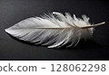 Single white feather lying on a dark gray surface 128062298