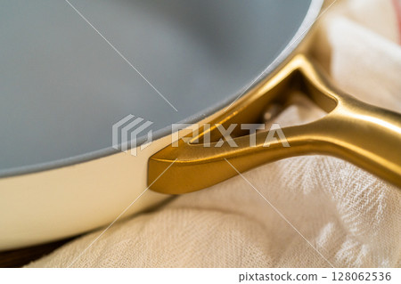 Side angle view of a ceramic skillet showing the junction of its smooth gray body and golden metallic handle, placed on soft white kitchen cloth. Image captured with shallow depth of field and warm 128062536