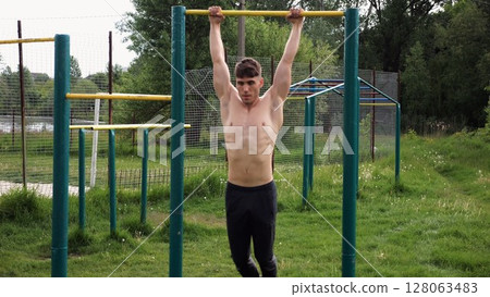 Muscular athlete doing pull ups on horizontal bar at sports ground. Young shirtless sportsman exercising at nature. Strong sporty man training outdoor. Concept of healthy and active lifestyle. Slow mo Muscular athlete doing pull ups on horizontal bar at sports ground. Young shirtless sportsman exercising at nature. Strong sporty man training outdoor. Concept of healthy and active lifestyle. Slow mo 128063483