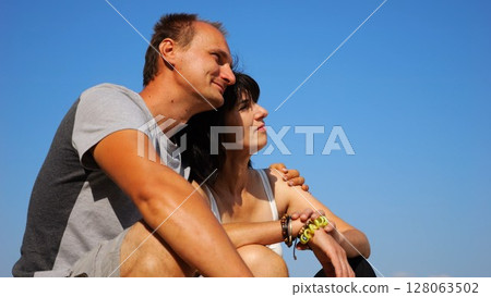 Happy pair hugs during sitting on wooden log on beach. Romantic couple admiring sea landscape at sunny day. Lovers spending time together at resort. Concept of summer vacation or holiday. Slow mo Happy pair hugs during sitting on wooden log on beach. Romantic couple admiring sea landscape at sunny day. Lovers spending time together at resort. Concept of summer vacation or holiday. Slow mo 128063502