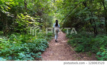 Young woman going along path at summer forest. Female with backpack walking among trail at wild nature. Unrecognizable girl enjoying stroll outdoor admiring beautiful scenic. Close up Rear view Young woman going along path at summer forest. Female with backpack walking among trail at wild nature. Unrecognizable girl enjoying stroll outdoor admiring beautiful scenic. Close up Rear view 128063520