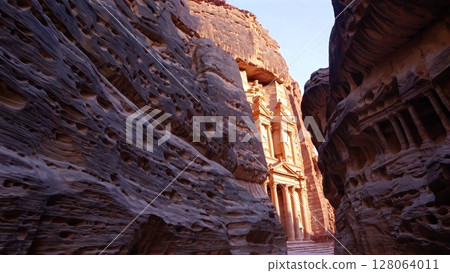 Warm sunset rays casting golden glow through narrow Siq canyon onto Al Khazneh Treasury, iconic sandstone monument in archaeological site of Petra, Jordan 128064011