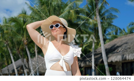 Young woman in white swimwear and straw hat walks along tropical beach Young woman in white swimwear and straw hat walks along tropical beach 128064474
