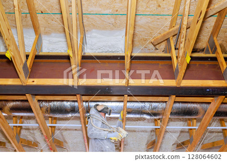 Worker in protective gear installs spray foam insulation with in exposed thermal barrier wooden framing at residential building. 128064602