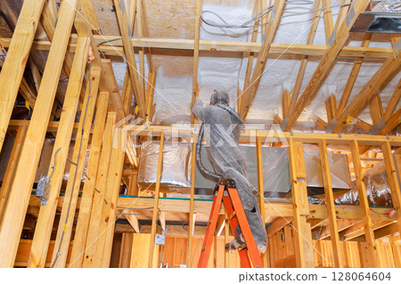Worker uses spray foam application to insulate ceiling beams in residential construction works 128064604