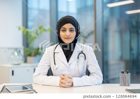 A female doctor wearing a hijab and headset, sitting at her desk, ready for a telemedicine consultation. A female doctor wearing a hijab and headset, sitting at her desk, ready for a telemedicine consultation. 128064694