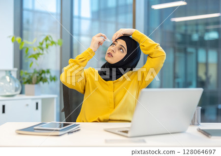 A businesswoman wearing a hijab applies eye drops in a modern office setting, likely to relieve eye strain while working on a laptop. 128064697